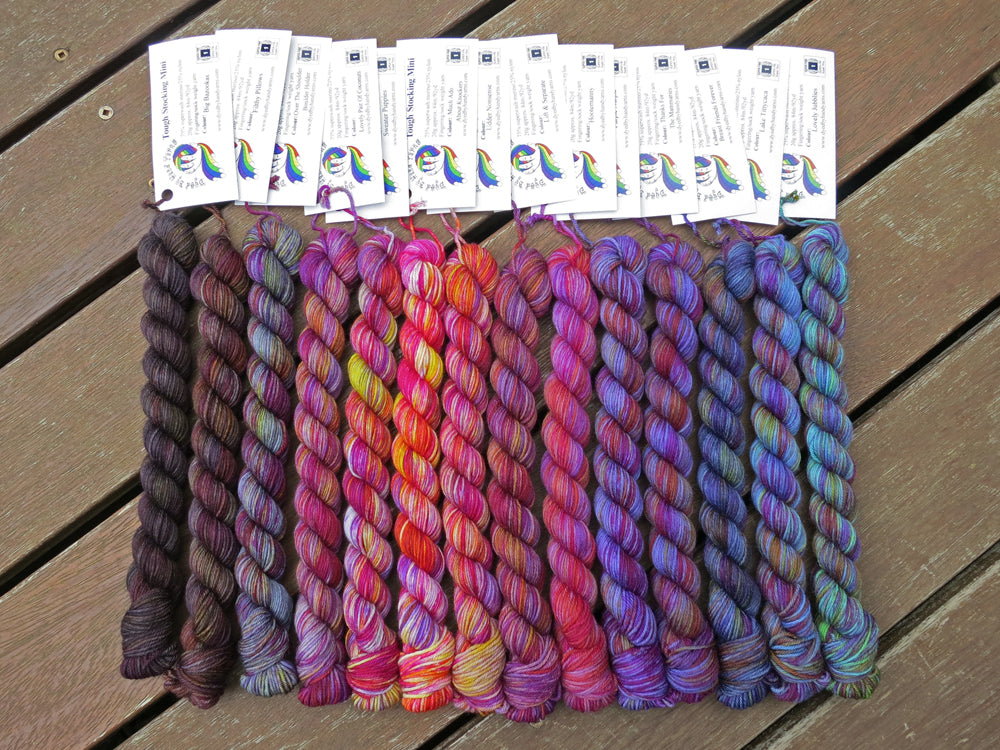 Fourteen variegated mini skeins of yarn in shades of grey, mauve and purple to blue with white labels are arranged from darkest grey at left to lightest grey then through mauve to purple then blues at right, lying on a brown wooden background.