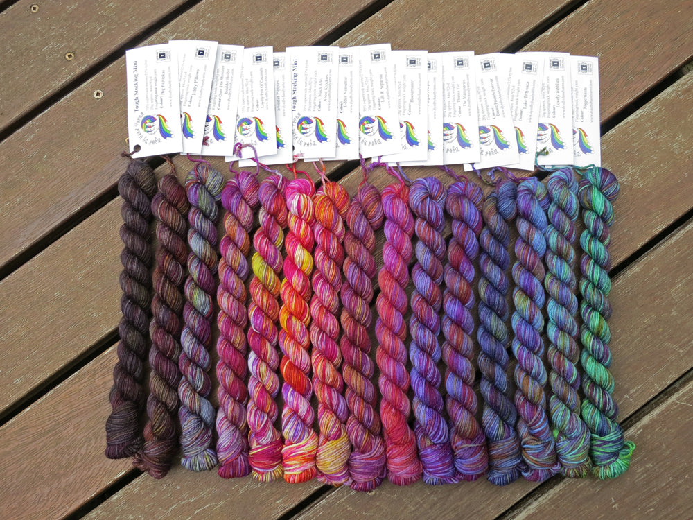 Fifteen variegated mini skeins of yarn in shades of grey, mauve and purple through blue and teal with white labels are arranged from darkest grey at left to lightest grey then through mauve and purple, blues then teal at right, lying on a brown wooden background.