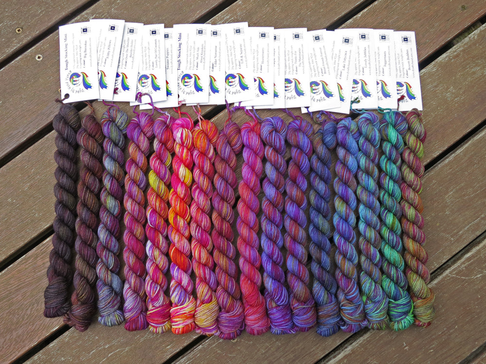 Seventeen variegated mini skeins of yarn in shades of grey, mauve and purple through blues and greens with white labels are arranged from darkest grey at left to lightest grey then through mauve and purple, blues then greens at right, lying on a brown wooden background.