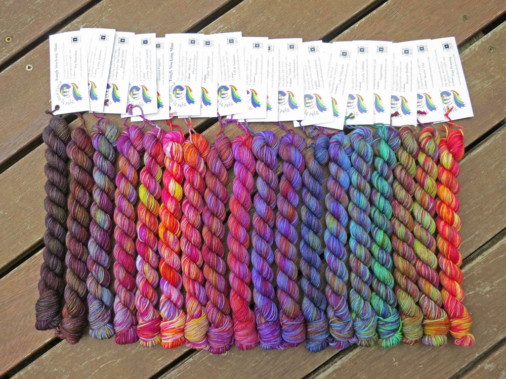 Eighteen variegated mini skeins of yarn in shades of grey, purple through blues, greens and yellow with white labels are arranged from darkest grey at left to lightest grey then through mauve and purple, blues, greens then yellow at right, lying on a brown wooden background.