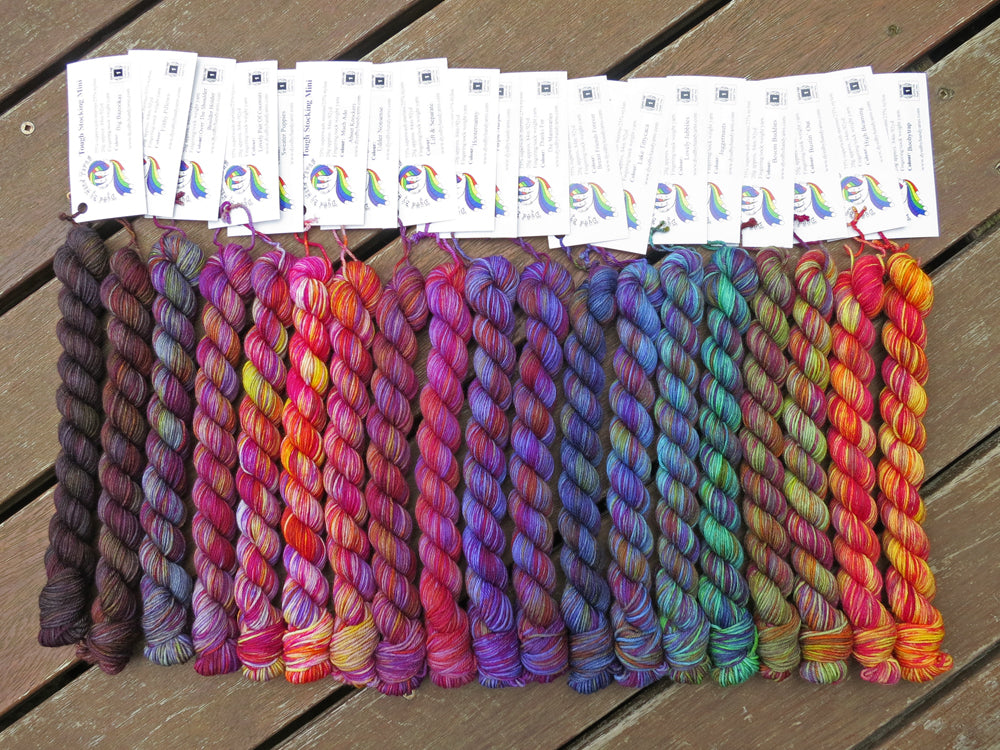 Nineteen variegated mini skeins of yarn with white labels are arranged from darkest grey at left to lightest grey then through purples, blues, greens and yellows at right, lying on a brown wooden background.