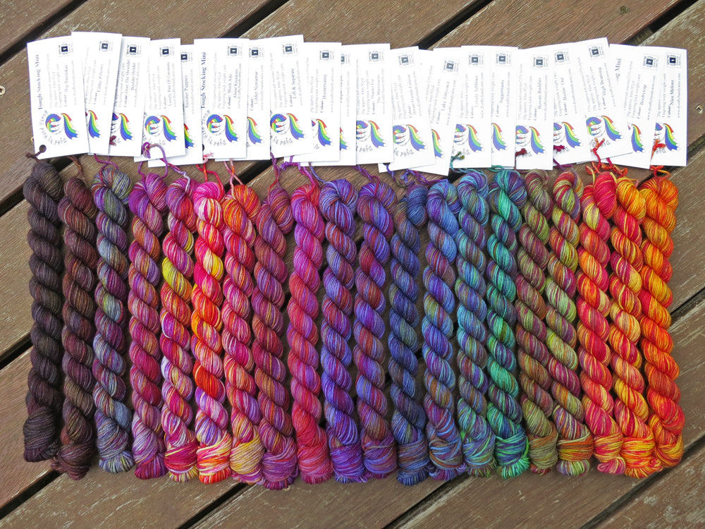 Twenty variegated mini skeins of yarn with white labels are arranged from darkest grey at left to lightest grey then through purples, blues, greens, yellows and orange at right, lying on a brown wooden background.