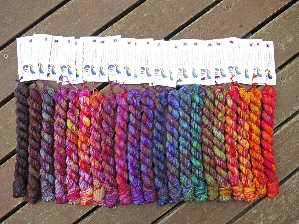 Twenty one variegated mini skeins of yarn with white labels are arranged from darkest grey at left to lightest grey then through purples, blues, greens, yellows and oranges at right, lying on a brown wooden background.