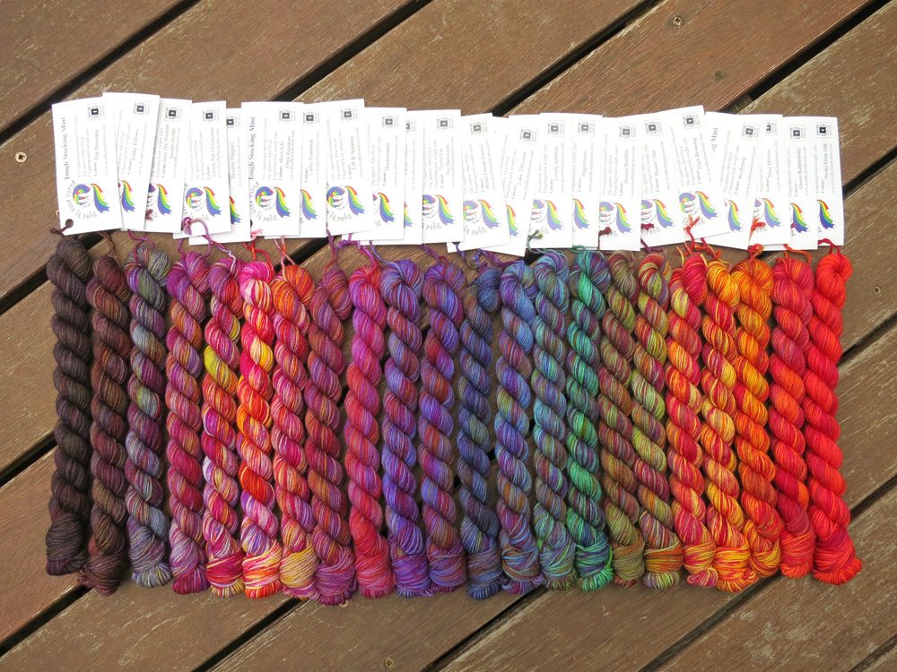 Twenty two variegated mini skeins of yarn with white labels are arranged from darkest grey at left to lightest grey then through purples, blues, greens, yellows, oranges and red at right, lying on a brown wooden background.