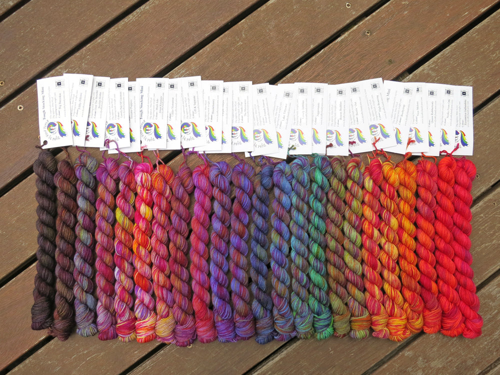Twenty three variegated mini skeins of yarn with white labels are arranged from darkest grey at left to lightest grey then through purples, blues, greens, yellows, oranges and reds at right, lying on a brown wooden background.