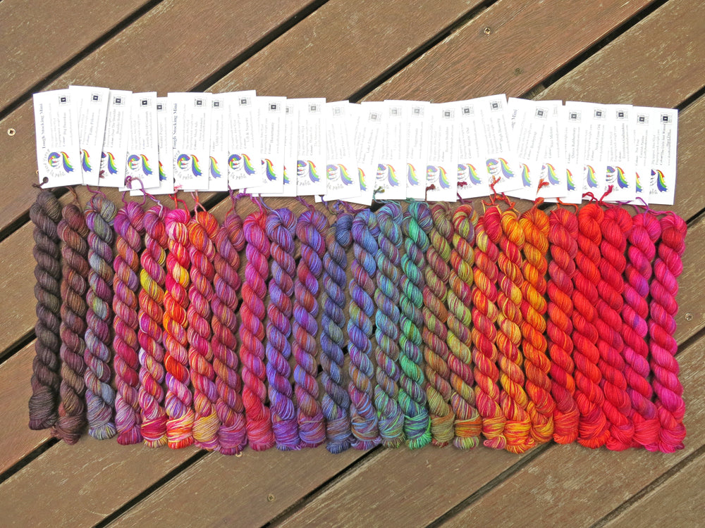 Twenty five variegated mini skeins of yarn with white labels are arranged from darkest grey at left to lightest grey then through purples, blues, greens, yellows, oranges, reds and pinks at right, lying on a brown wooden background.