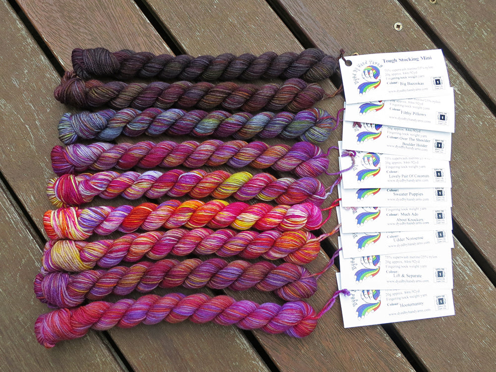 Nine variegated mini skeins of yarn in shades of grey and purple with white labels are arranged from darkest grey at top to lightest grey then to purple at bottom, lying on a brown wooden background.