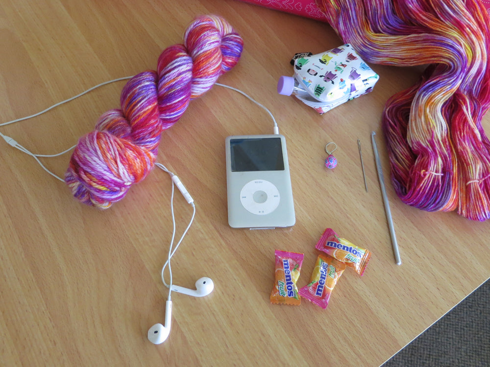 A bright variegated yarn skein of pink, purple, orange, yellow and white lies on a pale wooden background, next to an iPod and headphones, and various crafting tools