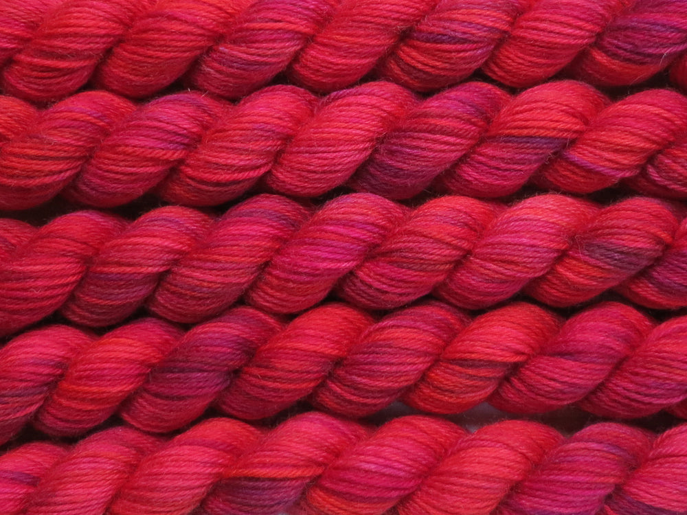 Multiple variegated mini skeins of yarn in crimson red with undertones of pink, purple, orange and yellow lying vertically on the screen.