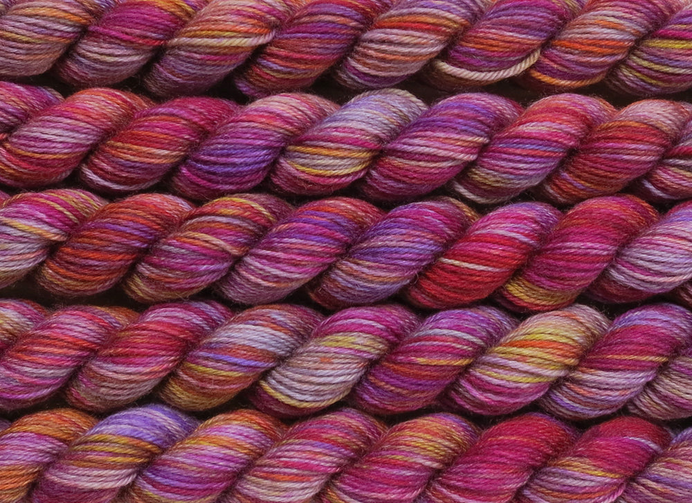 Multiple variegated mini skeins of yarn in light grey with undertones of pink, purple, orange and yellow lying vertically on the screen.