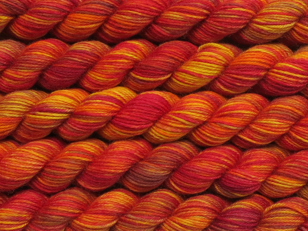 Multiple variegated mini skeins of yarn in golden orange with undertones of pink, purple, orange and yellow lying vertically on the screen.