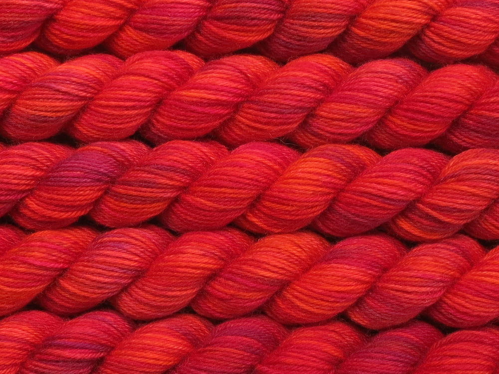 Multiple variegated mini skeins of yarn in orange red with undertones of pink, purple, orange and yellow lying vertically on the screen.
