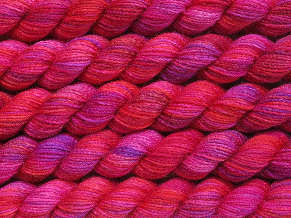 Multiple variegated mini skeins of yarn in magenta with undertones of pink, purple, orange and yellow lying vertically on the screen.