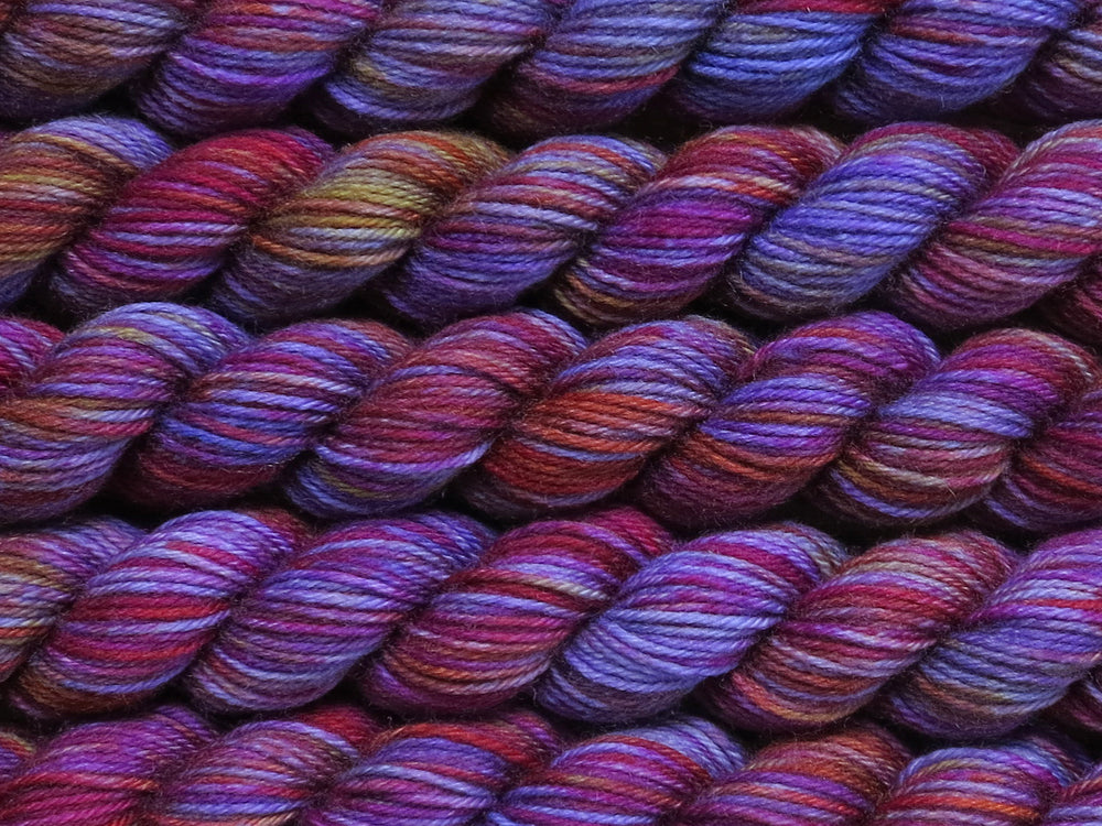 Multiple variegated mini skeins of yarn in deep purple with undertones of pink, purple, orange and yellow lying vertically on the screen.