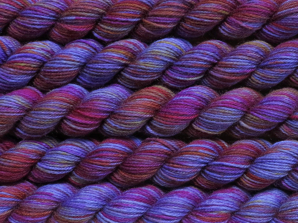 Multiple variegated mini skeins of yarn in violet blue with undertones of pink, orange and yellow lying vertically on the screen.