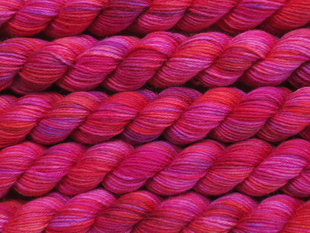 Multiple variegated mini skeins of yarn in fuchsia with undertones of pink, purple, orange and yellow lying vertically on the screen.