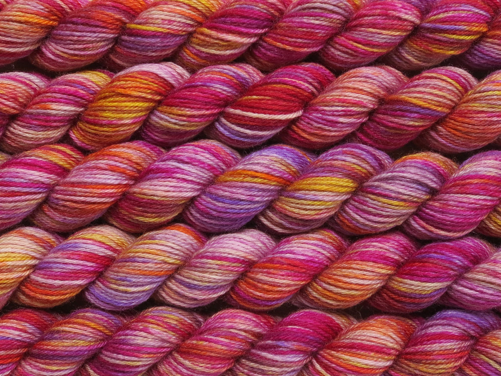 Multiple variegated mini skeins of yarn in pale mauve with undertones of pink, purple, orange and yellow lying vertically on the screen.