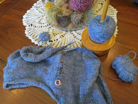 A piece of knitting in blue yarn with a dropped stitch held by a pink stitch marker. The knitting and three small ball of yarn lie on a brown wooden surface with a white crochet doily in the background.