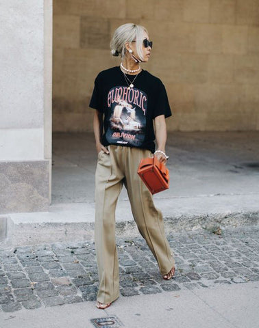 Woman walking on the street wearing a black punk chic graphic t-shirt, brown trousers and orange purse. 