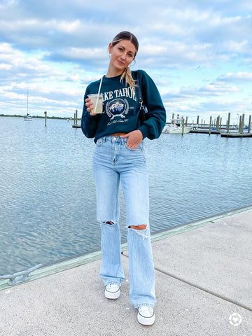 Stylish young woman wearing a blue sweatshirt, blue jeans and white shoes, standing next to a lake.