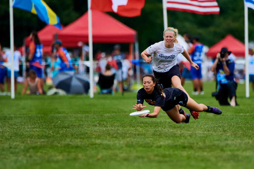 Two ultimate frisbee athletes competing at the 2022 WUCC. One athlete is doing a layout on the grass. Both are wearing VC Ultimate uniforms.