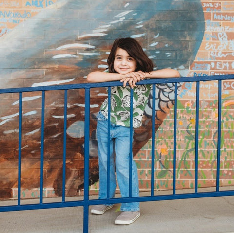 Young child with long brown hair, wearing jeans and a button up shirt stands behind dark, metal fence in front of painted wall mural. 