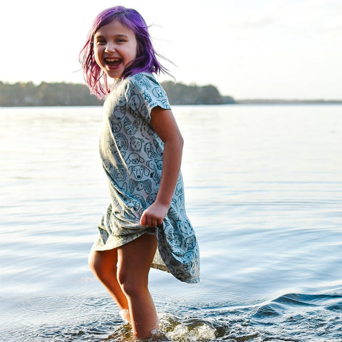 Child with purple hair in blue dress with dogs printed on it stands up to her shins in a lake