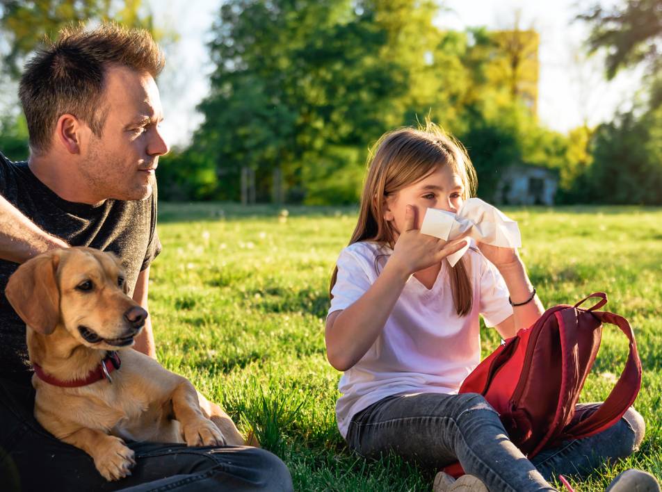 Et barn sidder på en græsplæne med en mand og en hund. Barnet tørrer næse med en serviet.