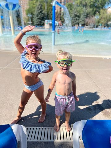 Boy and girl toddlers striking a pose while wearing Frogglez Goggles Navigatorz Half Mask goggles. Standing in front of a water splash park.