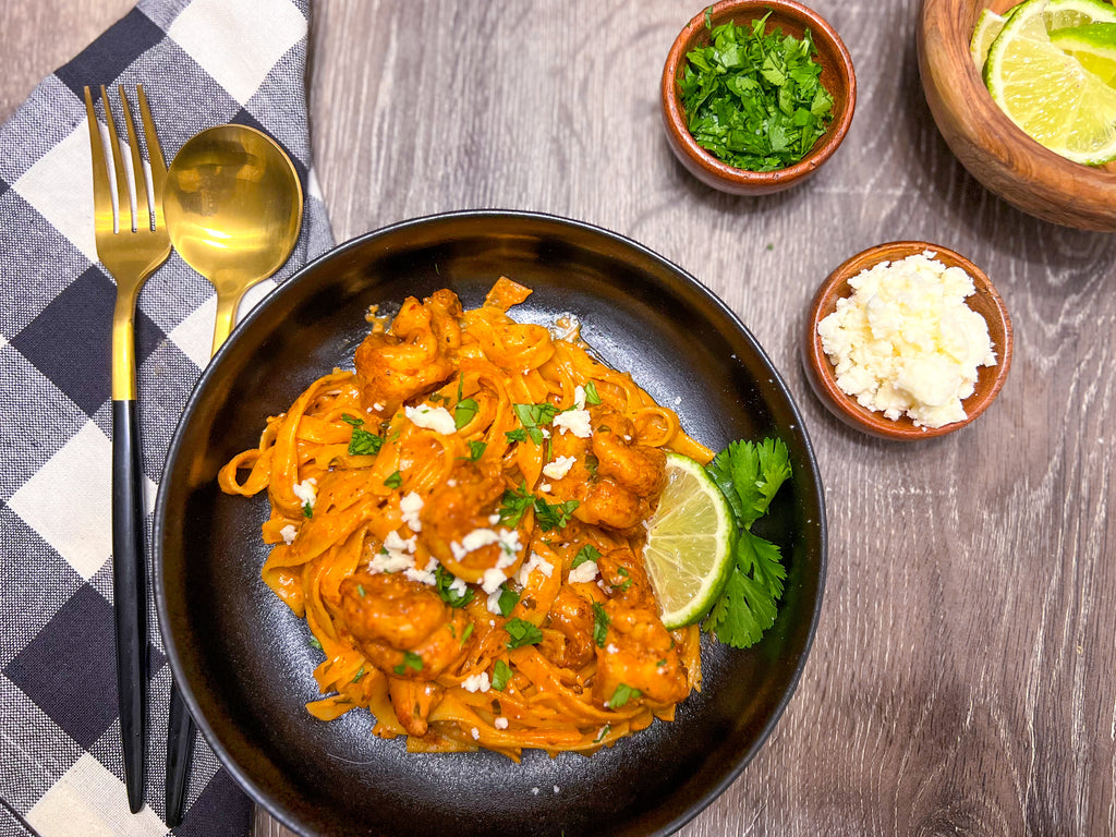 creamy guajillo tequila lime pasta with rojo smoke shrimp flatlay in a black flat bowl with gold and black utensils and ramekins of lime wedges queso fresco and chopped cilantro