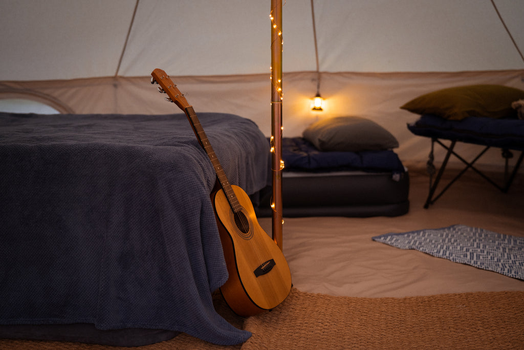 Una guitarra se apoya en el pie de un colchón de aire tamaño queen cerca del poste central de una tienda de campaña de lona, ​​con dos camas individuales al fondo.