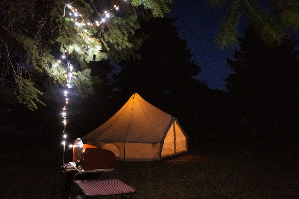 Una serie de luces parpadeantes iluminan el primer plano con una cocina de campamento debajo de los árboles, y una tienda de campaña de lona se ilumina desde el fondo.
