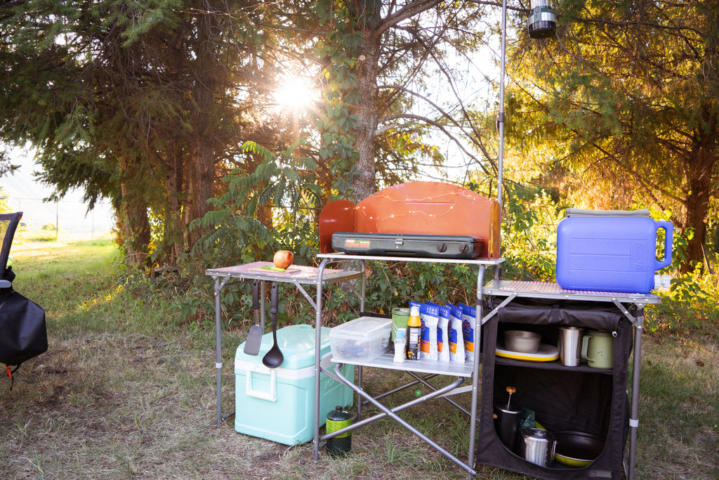 Una cocina al aire libre que incluye una hielera, una estufa para acampar, una jarra de agua portátil y varios suministros de cocina para acampar.