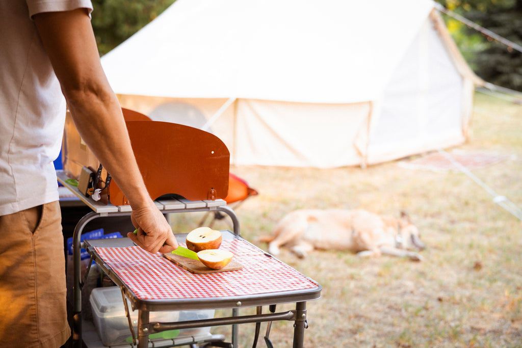 Una persona se encuentra detrás de una cocina de campamento al aire libre cortando una manzana, mientras un perro duerme a lo lejos frente a una tienda de campaña de lona.
