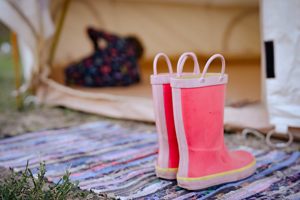 Une paire de bottes de pluie roses repose sur un tapis tressé coloré devant les portes ouvertes d’une tente cloche en toile.