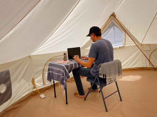 Un bureau est installé à l’intérieur d’une tente polyvalente en toile avec un cordon d’alimentation passant par le port d’accès et un homme est assis au bureau devant un ordinateur portable.