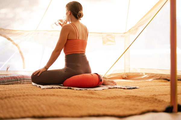 Une femme en tenue de yoga est assise sur un coussin de méditation, sirotant de l'eau et regardant les murs grillagés d'une tente polyvalente en toile.