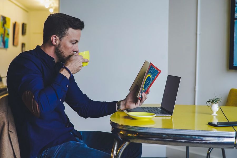 Man reading a book and checking his laptop while enjoying his CBD coffee