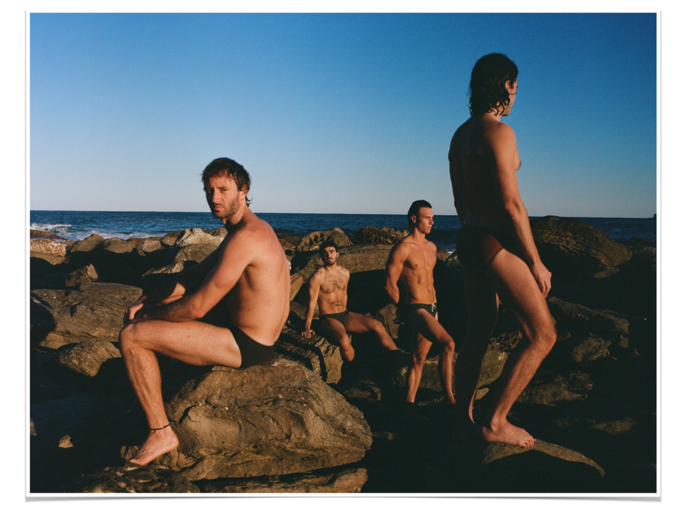 Men sitting and standing staggered on rocks near beach.