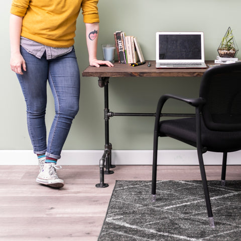 An image of a person standing by a desk organized with PIPE DECOR®, featuring a laptop, books, and a plant on a wooden surface in a stylish workspace.