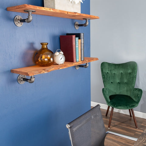 Image of a floating shelf by PIPE DECOR®, mounted on a blue wall with vintage books, a glass vase, and an alarm clock, next to a green chair.