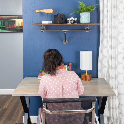 Image of a woman seated at an industrial-style desk by PIPE DECOR®, featuring vintage cameras and a plant on a floating shelf against a vibrant blue wall, showcasing a stylish and functional workspace.