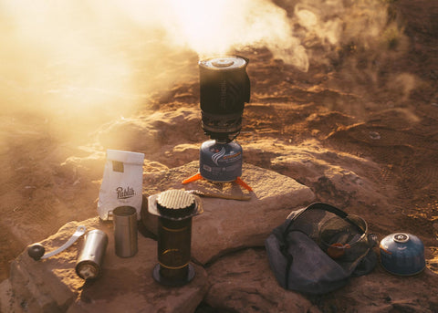 Supplies for making coffee set up on ground: A bag of coffee, grinder, Jet Boil, and fuel. Water is steaming.