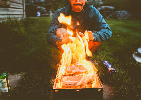 Man squatting in front of grill, cooking steaks over roaring fire.