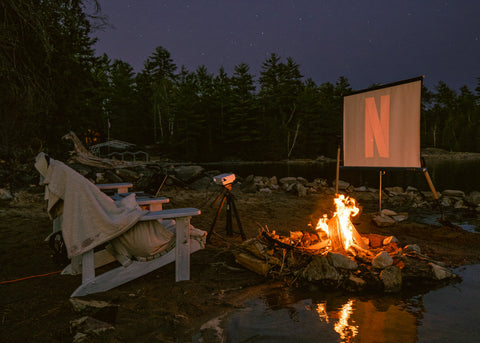 Outdoor movie setup with projector, a few chairs, and a campfire with forest backdrop.