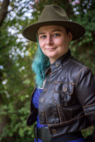 A woman is standing amongst greenery and fallen leaves, wearing a purple skater dress with black cats on it, tight black pants, knee-high black boots, a black waist belt, a dark brown cropped faux leather jacket, and an olive green wide brimmed hat.