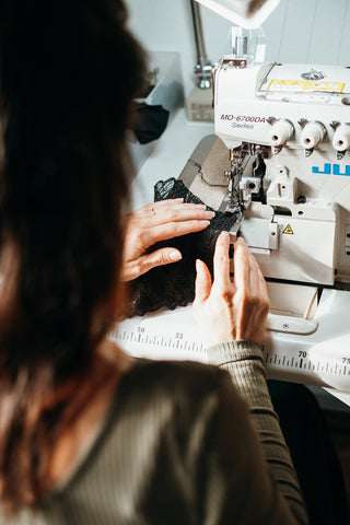 Foto zeigt unser Teammitglied Kira an der Overlock Nähmaschine. Die Perspektive des Bildes ist von hinten, es sieht aus, als würden wir ihr über die Schulter sehen. Zartes Abendlicht fällt von der Seite auf die Nähmaschine.