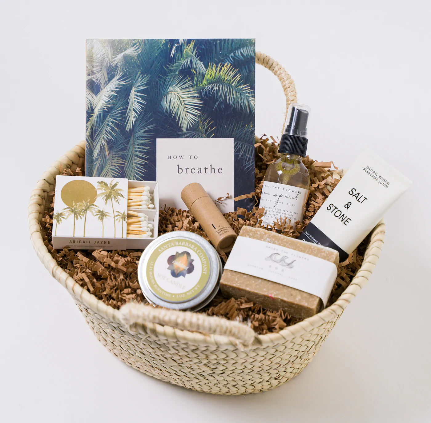 A basket filled with a book, a candle, matches, lip balm, a spray, sunscreen, and soap