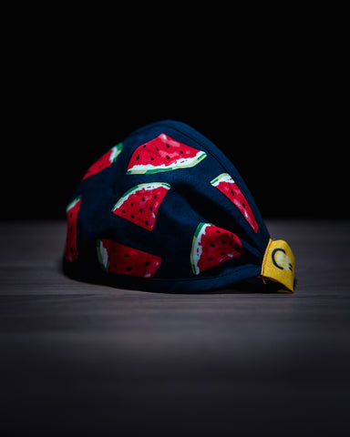 Blue facemask with red watermelon wedges on it on a wooden boar in front of a jet black background