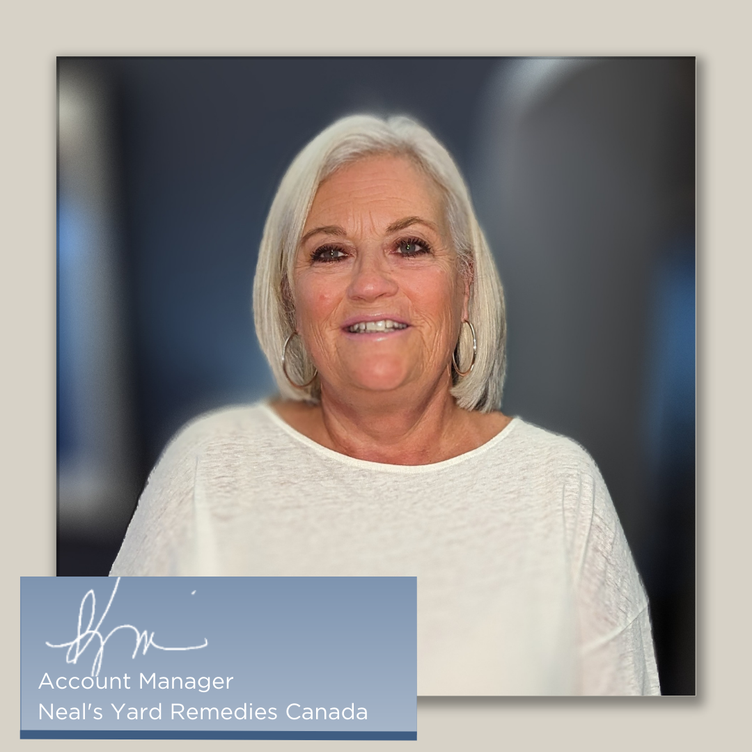 A mature woman stands confidently, wearing a cream long sleeve shirt. She is standing in front of blurred background, with a sign stating role in Neals Yard Remedies as a Account Manager. Her friendly smile and welcoming demeanor invite visitors to step inside and explore the world of natural and organic skin care products.